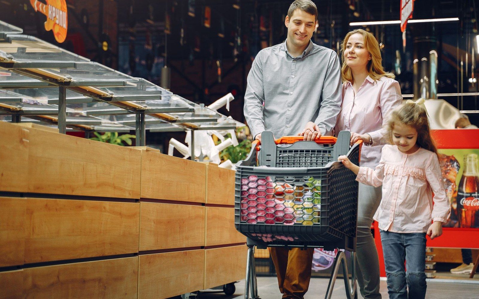 Parents and their young daughter push a full shopping cart through a grocery store aisle