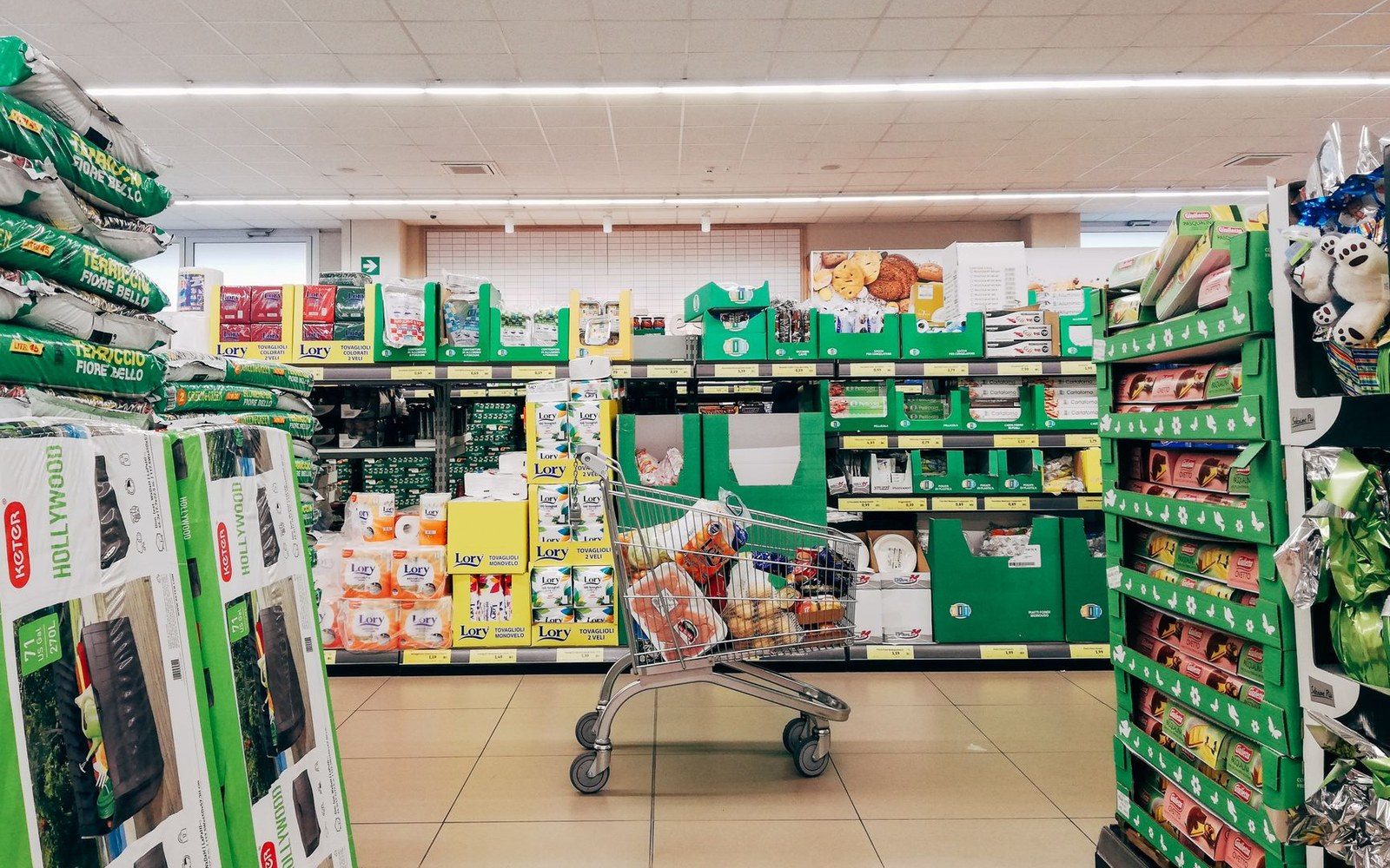 A shopping cart with groceries sits in a supermarket aisle stocked with green-packaged goods